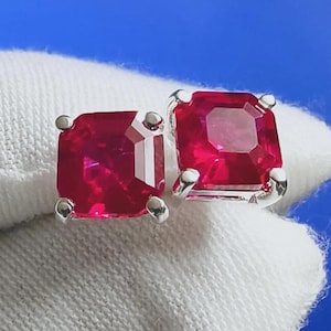 May include: A pair of square-cut, ruby-red gemstone stud earrings. Each earring features a vibrant, faceted red stone set in a silver-colored metal setting. The earrings are displayed on a white fabric background with a blue backdrop.