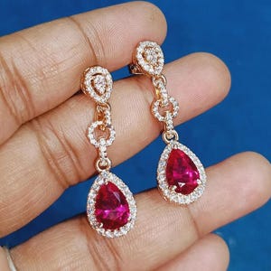 May include: A pair of rose gold earrings with a teardrop-shaped, ruby-red gemstone. The earrings feature a halo of small, clear stones around the ruby-red gemstone and at the top of the earring. The earrings are connected by a series of small, clear stones.