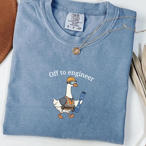 May include: A blue Comfort Colors t-shirt with the phrase "Off to engineer" above a cartoon goose wearing a hard hat, safety glasses, and tool belt, holding a blueprint. A gold necklace is draped across the shirt.