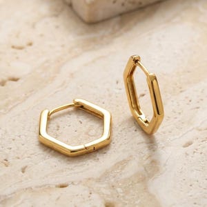 Puede incluir: Dos pendientes de aro geométricos dorados. Cada pendiente tiene forma hexagonal. Los pendientes se muestran sobre una superficie clara y texturizada. Los pendientes son de metal brillante.