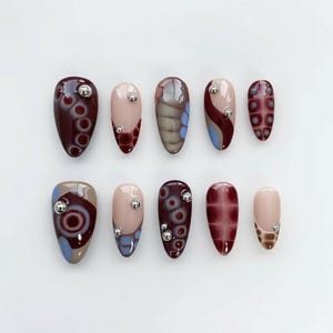 Può includere: Un set di dieci unghie finte con vari design. Le unghie presentano un mix di colori, tra cui bordeaux, beige e blu, con motivi decorativi e abbellimenti argentati. Le forme delle unghie sono ovali e a mandorla.