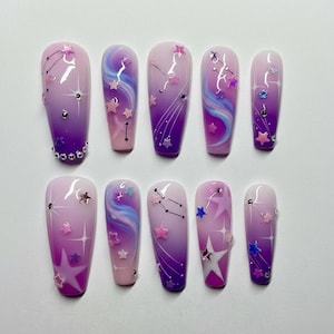 May include: A set of ten long, coffin-shaped false nails. Each nail features a gradient of purple and pink with star and constellation designs. Some nails have silver accents and rhinestones.