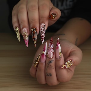 May include: A set of long, pointed acrylic nails with intricate designs. The nails feature gold accents, floral patterns, and a mix of colors including red, white, pink, and brown. Some nails have a textured, gold finish.