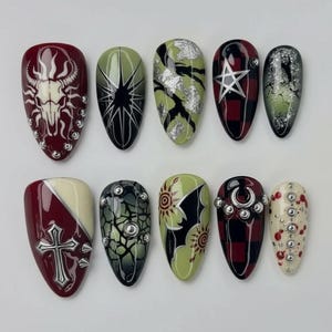 Puede incluir: Un conjunto de diez uñas postizas con varios diseños. Las uñas presentan una mezcla de colores rojo oscuro, verde y negro con adornos como tachuelas plateadas, cruces y patrones de estrellas. El arte de uñas incluye diseños de calaveras, florales y geométricos.