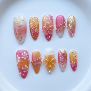 Op de afbeelding: Een set van tien kunstnagels met een tropisch thema. De nagels hebben verschillende ontwerpen, waaronder gouden bloemaccenten, ombre-effecten en 3D-bloemversieringen. Kleuren zijn wit, roze, oranje en goud.