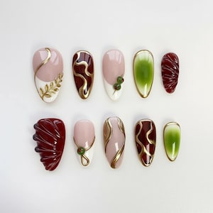 May include: A collection of ten artificial nails with various designs. The nails feature a combination of colors, including pink, white, green, and burgundy, with gold accents. Some nails have leaf and swirl patterns.