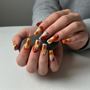 May include: Long, square-shaped acrylic nails with a colorful abstract design in shades of orange, yellow, and white. Some nails feature small silver embellishments. The nails are painted with a glossy finish.