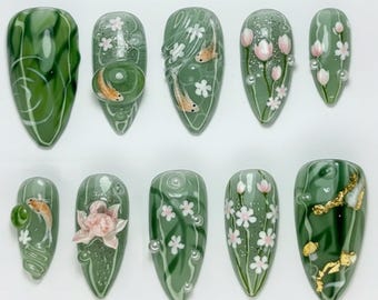 May include: A set of ten almond-shaped, green press-on nails with various designs. Some nails feature koi fish, pink flowers, and white accents. Others have a marbled effect or gold leaf details, creating a unique and artistic manicure.