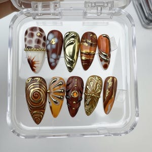 May include: A collection of ten false nails displayed in a clear case. The nails showcase diverse designs, including gold embellishments, swirls, and patterns in brown, orange, and white. Some nails have a textured, three-dimensional appearance.
