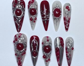 Puede incluir: Un conjunto de diez uñas artificiales con varios diseños. Cada uña presenta un color base rojo intenso, adornado con adornos plateados y una gema roja grande y redonda. Algunas uñas tienen una base blanca con detalles rojos y plateados.