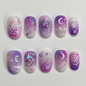 Puede incluir: Un conjunto de diez uñas artificiales con temática espacial. Cada uña presenta un diseño diferente, incluyendo estrellas, lunas, planetas y galaxias arremolinadas, en tonos morados, rosas y blancos. Algunas uñas tienen purpurina y pequeños adornos.