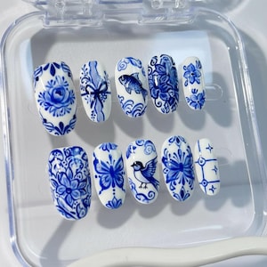 Puede incluir: Un conjunto de diez uñas postizas blancas con intrincados diseños florales y animales azules. Los diseños incluyen flores, un pez, un pájaro y patrones de lazo. Las uñas son de forma ovalada y se muestran en una caja de plástico transparente.