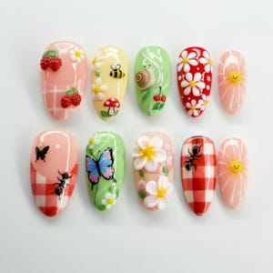 Puede incluir: Un conjunto de diez uñas artificiales con varios diseños. Las uñas presentan ilustraciones detalladas que incluyen cerezas, abejas, caracoles, margaritas, mariposas, hormigas y soles sonrientes. Los colores incluyen rosa, amarillo, verde y rojo, con un patrón de cuadros en algunos.