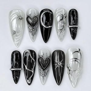 May include: A set of ten long, pointed artificial nails. Designs include black and silver hearts, moons, and abstract patterns. Some nails feature metallic accents, creating a gothic and edgy aesthetic. The color palette is primarily black, white, and silver.
