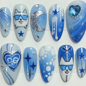 Könnte beinhalten: Ein Set aus zehn blauen und weißen Kunstnägeln mit verschiedenen Designs. Die Designs umfassen einen Hund mit Sonnenbrille, einen Mond, Sterne und einen herzförmigen Edelstein. Einige Nägel haben Glitzerakzente.