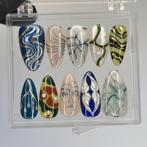 May include: A set of ten artificial nails in a clear plastic case. Each nail features a unique design, including abstract patterns, geometric shapes, and facial art. Colors include blue, beige, green, and gold, with silver and white accents.