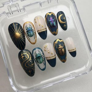May include: A set of ten artificial nails with various designs. The nails feature dark blue, white, and gold accents. Some nails have beetle illustrations, while others display celestial patterns like stars and moons. The nails are oval-shaped and presented in a clear plastic case.