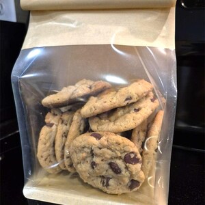 May include: A clear plastic bag with a brown paper top filled with chocolate chip cookies. The cookies are golden brown with dark chocolate chips. The bag is sealed and sits on a dark surface.