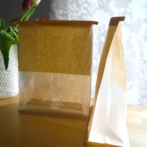 May include: Two brown paper bags, one upright and one folded, sit on a wooden surface. The bags have a clear, transparent section at the bottom. A vase with tulips is in the background, with a floral patterned wallpaper.