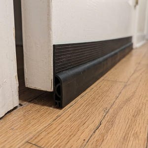 May include: A black rubber door sweep attached to the bottom edge of a white door. The door sweep features a ribbed upper section and a hollow, flexible lower seal that rests against the floor to block gaps beneath the door frame.