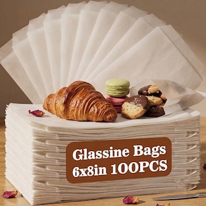 May include: A large stack of translucent white glassine bags with a brown rectangular label reading Glassine Bags 6x8in 100PCS. A golden brown croissant, a green macaron, a pink macaron, and several chocolate chip cookies are placed on top.