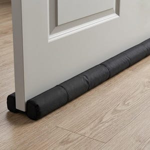 May include: A black fabric door draft stopper positioned at the base of a white interior door. The cylindrical, padded accessory features a segmented design and fits snugly against the bottom edge of the door to cover the gap between the door and floor.