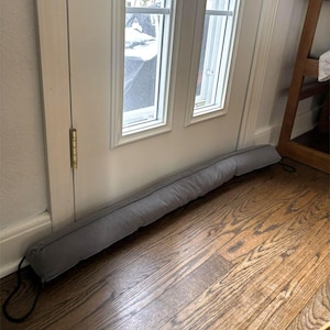 May include: A long, gray fabric door draft stopper rests on a hardwood floor. The cylindrical cushion is positioned at the base of a white door with a window. Black cords are attached to each end of the stopper.
