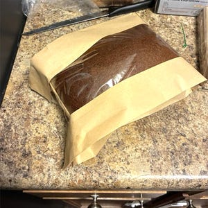 May include: A loaf of brown bread wrapped in clear plastic and brown paper. The bread is rectangular and appears freshly baked. The packaging is neatly folded, suggesting a bakery or homemade product.