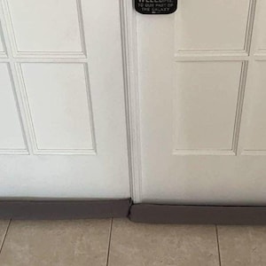 May include: Two white French doors with a gray draft stopper at the base. The doors have rectangular panels and are closed. The draft stopper is a long, cylindrical foam tube designed to block drafts and conserve energy.