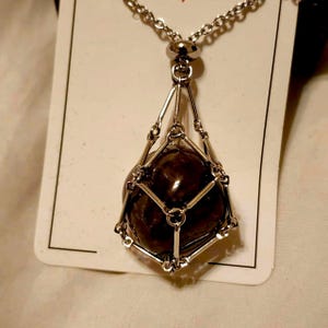 May include: A silver-toned necklace featuring a dark brown, orb-shaped pendant. The pendant is encased in a cage-like structure, suspended from a delicate chain. The pendant has a polished, reflective surface.