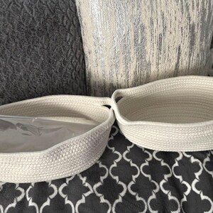 May include: Two oval-shaped off-white woven rope baskets with integrated handles. One basket contains a clear plastic bag inside, while the other is empty. The baskets feature a thick, coiled texture and are positioned side by side on a patterned surface.