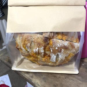 May include: A loaf of artisan bread, sliced and visible through a clear plastic window in a brown paper bag. The bread has a golden-brown crust with white flour dusting and visible air pockets.