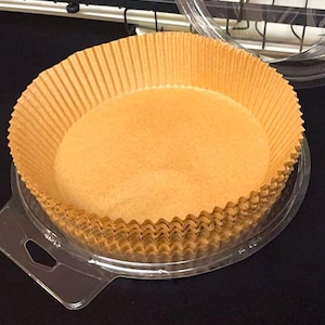 May include: Stack of brown, corrugated paper baking cups in a clear plastic container. The cups are round with a fluted edge. The container has a handle and is sitting on a black surface.