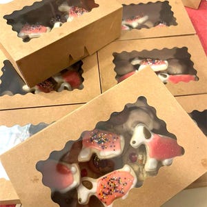 May include: Several brown cardboard boxes with clear window cutouts, each containing decorated cookies. The cookies are shaped like dogs and other shapes, with pink, white, and brown icing and colorful sprinkles.