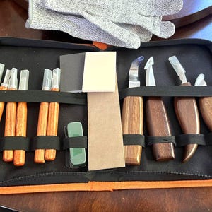 May include: A black zippered case with orange trim, containing wood carving tools. The set includes various knives, chisels, and gouges with wooden handles, along with protective gloves and sharpening stones. The tools are secured with black elastic straps.