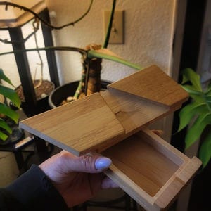 May include: A light-colored wooden box with a sliding top and a small drawer. The box is held open, revealing the interior. The wood grain is visible, and the box appears to be new.