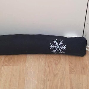 May include: A black fabric draft stopper with a white embroidered snowflake design. The long, cylindrical cushion rests on a wooden floor, near a white door and a teal wall.
