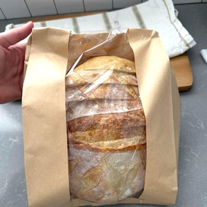 Peut inclure: Un pain fraîchement cuit, emballé dans un sac en papier brun. Le pain a une croûte brun doré et est partiellement visible à travers l'emballage plastique transparent. Le sac est tenu par une main.