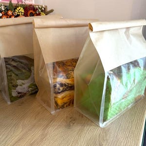 May include: Three loaves of bread are visible in the image, each packaged in a brown paper bag with a clear plastic window. The bread varieties include a green loaf, a marbled loaf, and a loaf with a golden crust. The bags are on a wooden surface.