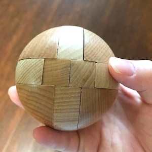 May include: A wooden puzzle ball composed of interlocking wooden pieces. The ball is a light brown color and appears to be made of multiple rectangular blocks of wood. The puzzle is held in a hand.