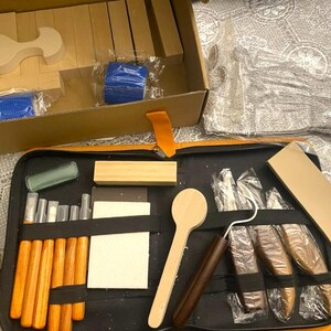 May include: A craft kit with various wooden tools and components. The kit includes a wooden spoon, carving tools, and rectangular and curved wooden blocks. The tools are arranged in a black zippered case with an orange handle, and a cardboard box.
