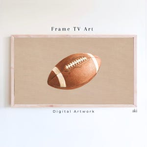 Op de afbeelding: Een digitaal kunstwerk in een lijst met een bruine American football met witte strepen en stiksels. Het kunstwerk staat tegen een beige achtergrond en is omlijst in licht hout. De tekst luidt "Frame TV Art" en "Digital Artwork".