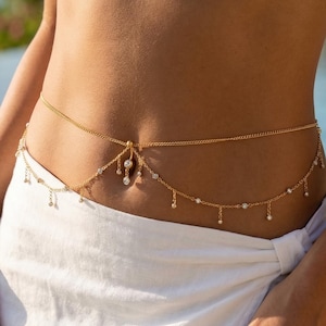 May include: A gold-colored body chain with dangling charms and small white beads. The chain is worn over a white skirt and top. The background includes a swimming pool and palm trees, suggesting a beach or resort setting.