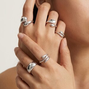 May include: A selection of silver rings in diverse organic forms. The rings are shown on a hand, highlighting their smooth, reflective surfaces. Designs include a dome, a wrap-around style, and teardrop shapes.