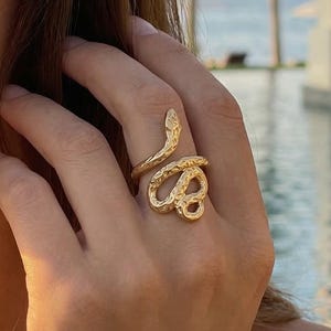 Puede incluir: Un anillo de serpiente dorado con detalles texturizados. El anillo se envuelve alrededor del dedo, con la cabeza y la cola de la serpiente formando los extremos. El anillo se lleva en una mano, con un fondo borroso de una piscina y un paisaje tropical.