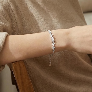 May include: A silver bracelet with a leaf design is worn on a person's wrist. The bracelet features small, clear stones. The person is wearing a beige sweater and holding a mug. The background includes a window and neutral-toned pillows.