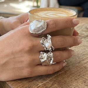 Puede incluir: Tres anillos de plata con diseños orgánicos y texturizados. Un anillo presenta una perla grande e irregular. Los otros dos anillos tienen formas abstractas. Los anillos se llevan en una mano que sostiene una taza de café. El entorno parece ser un café.