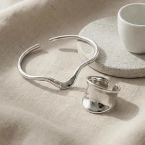 May include: A silver-toned cuff bracelet with a wavy design and an open-ended ring are displayed on a neutral-toned surface. A small white ceramic cup sits on a gray stone. The jewelry has a polished, reflective finish.