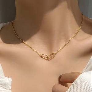Puede incluir: Un collar dorado con una cadena delicada y dos colgantes ovalados entrelazados. El collar se muestra en el cuello de una persona, mostrando su diseño. La joya es simple y elegante.