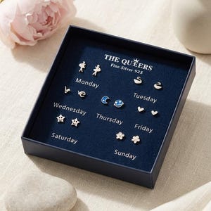 Op de afbeelding: Een set fijne zilveren oorbellen in een marineblauwe doos. De oorbellen hebben verschillende vormen, waaronder sterren, harten en bloemen. De doos is gemarkeerd met "THE QUEERS Fine Silver 925" en heeft de dagen van de week erop.